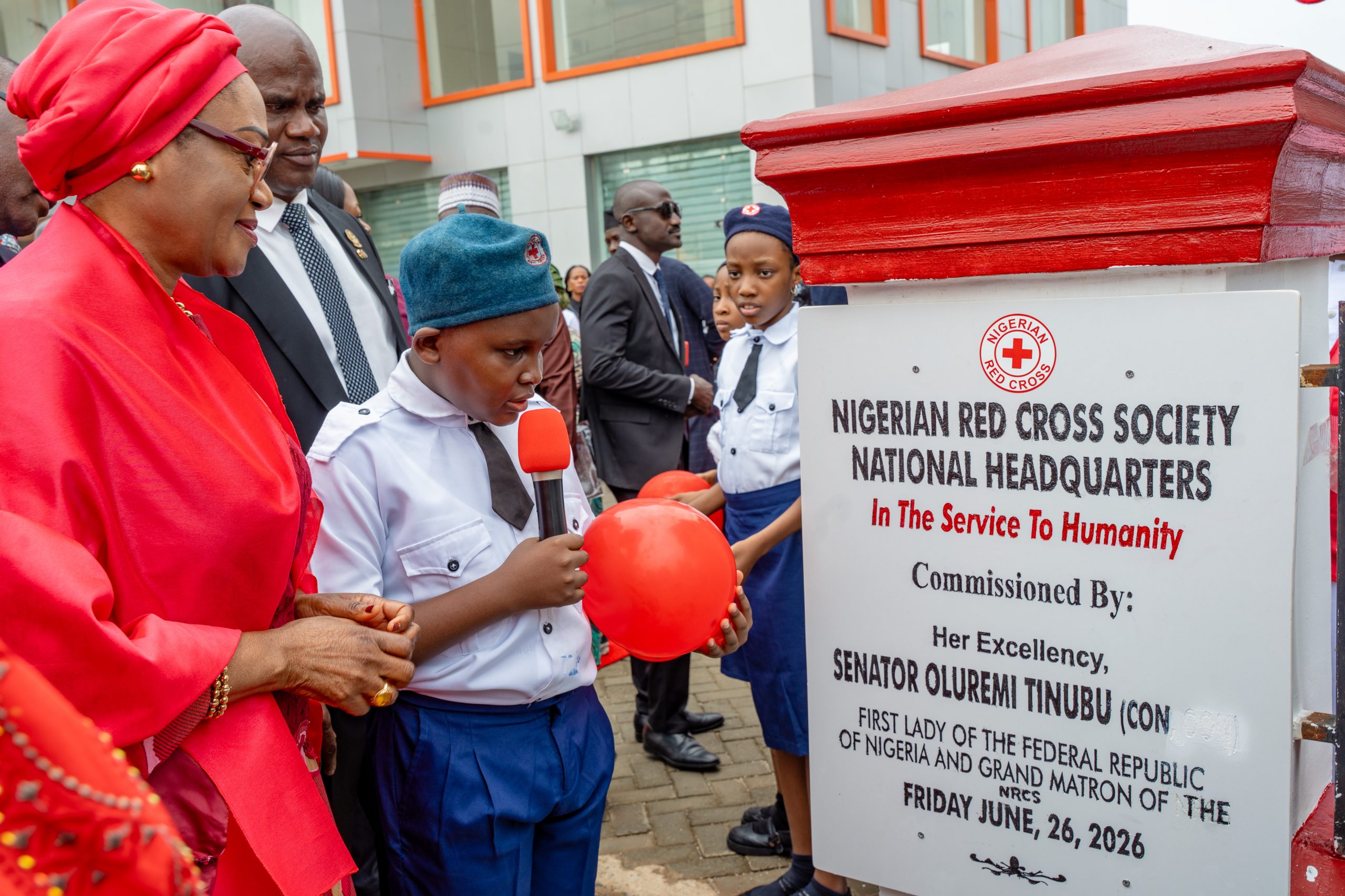 First Lady Senator Oluremi Tinubu, CON, looking at a commemorative plaque unveiled by a young boy holding a microphone and a red balloon.