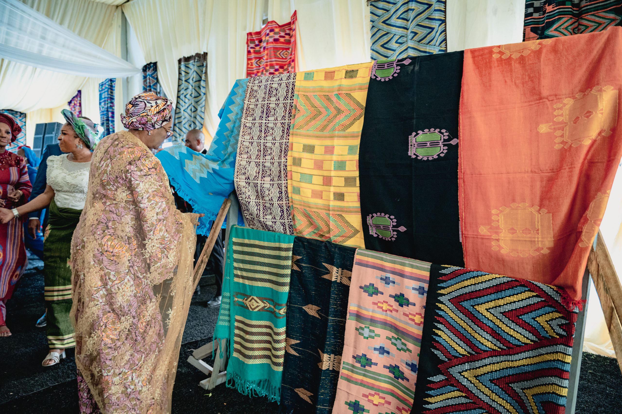 First Lady Senator Oluremi Tinubu, CON, wearing a patterned gold and pink outfit and head wrap, looking at colorful traditional fabrics displayed on a rack.