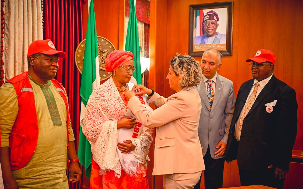 First Lady Senator Oluremi Tinubu, CON, being decorated with a sash and pin by an official, flanked by Red Cross representatives and dignitaries indoors.