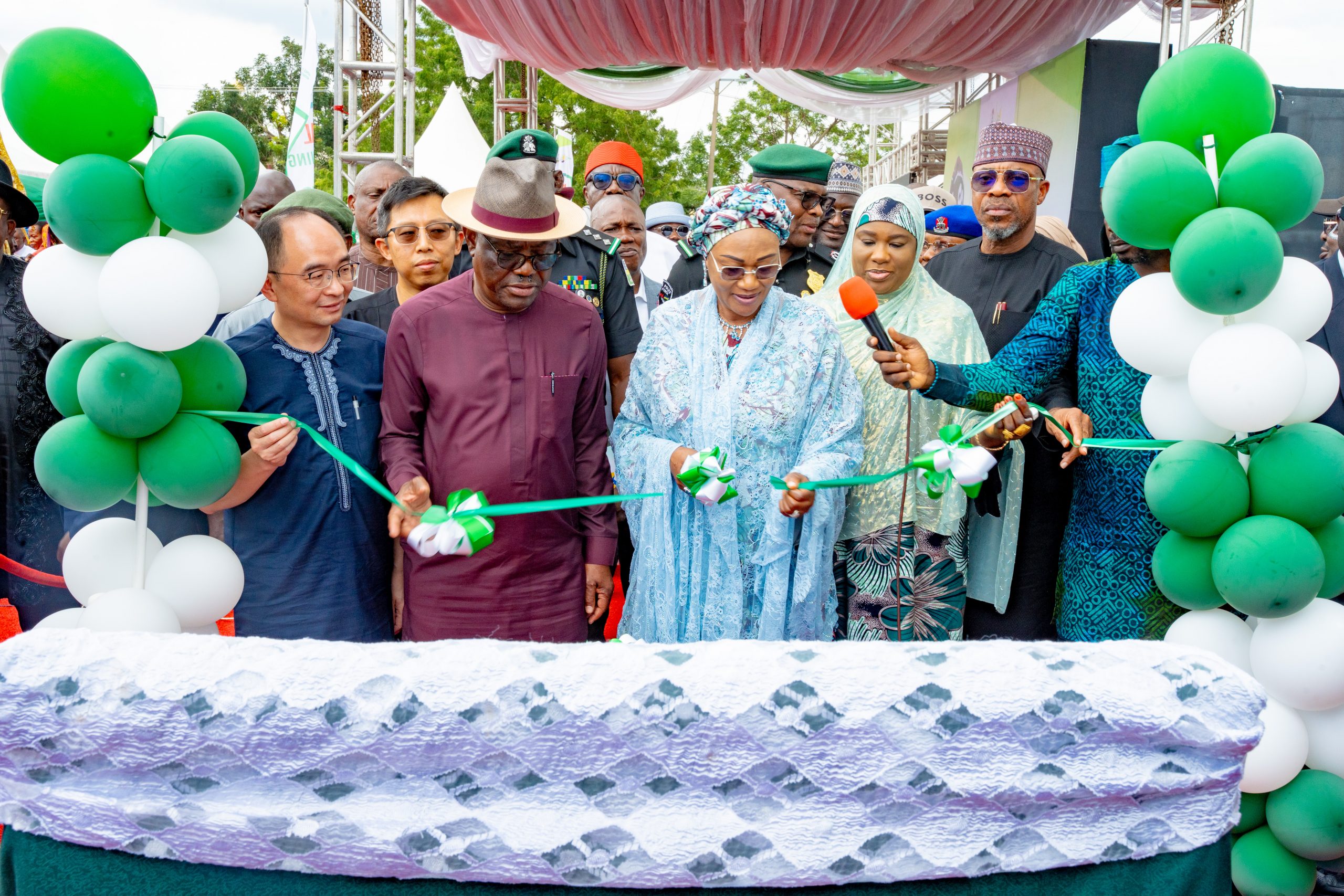 First Lady Senator Oluremi Tinubu, CON, cutting a ribbon alongside officials amidst green and white balloons during an outdoor commissioning ceremony.