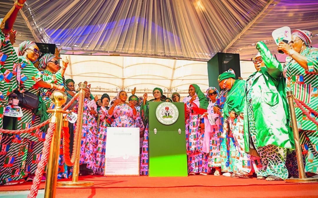 First Lady Senator Oluremi Tinubu, CON, standing behind a green podium bearing the Nigerian coat of arms, surrounded by cheering supporters.
