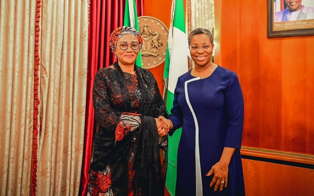First Lady Senator Oluremi Tinubu, CON, wearing a patterned dark red and black outfit with a matching head wrap, shaking hands with a woman in a blue dress indoors.
