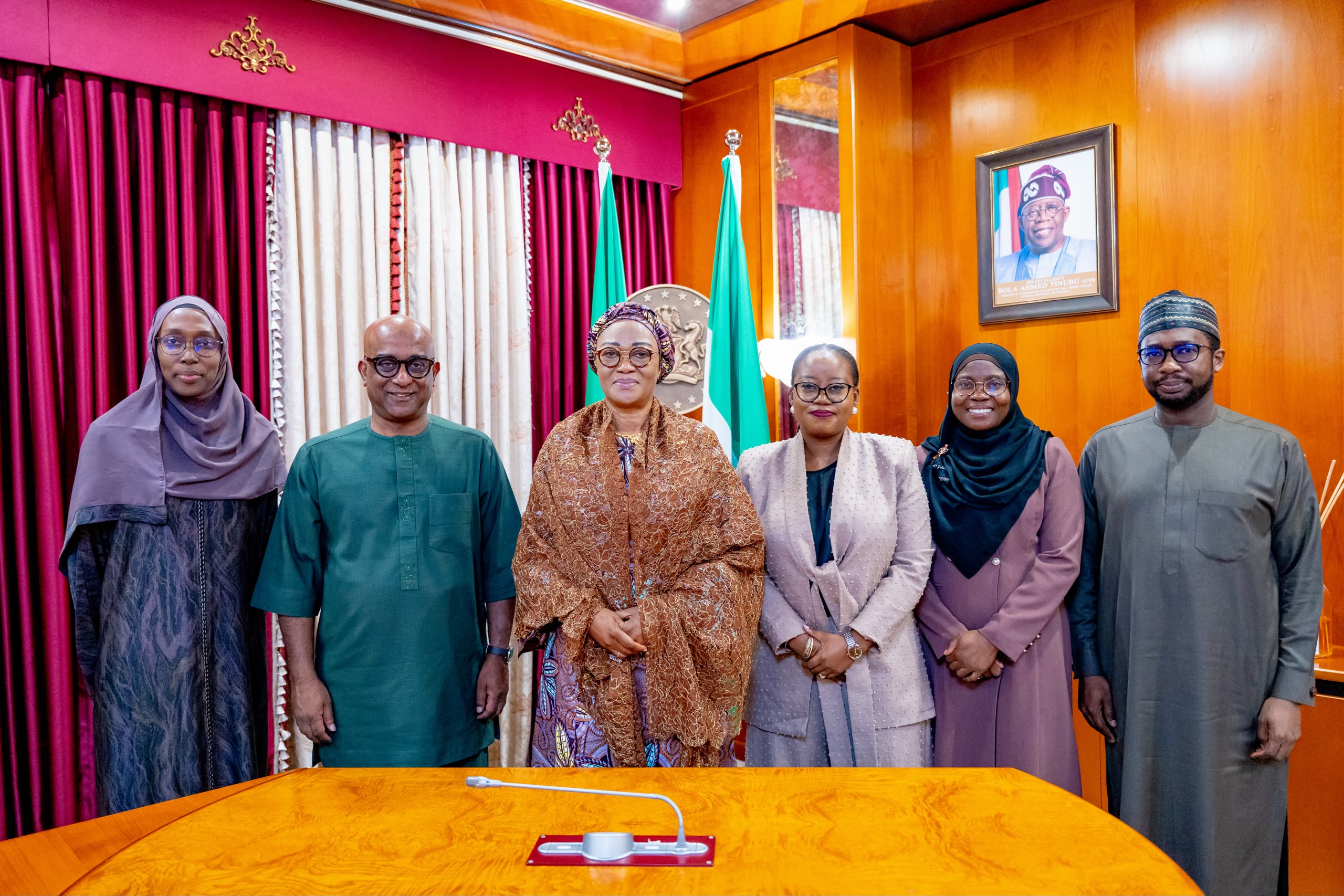 First Lady Senator Oluremi Tinubu, CON, wearing a patterned brown outfit and wrap, standing center behind a table alongside a group of officials and dignitaries indoors.