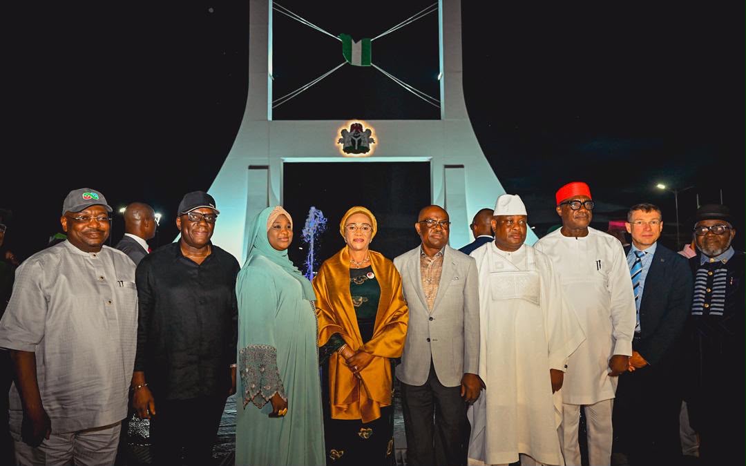 First Lady Senator Oluremi Tinubu, CON, wearing a dark outfit and gold wrap, standing with a group of government officials and dignitaries in front of a large illuminated monument at night.