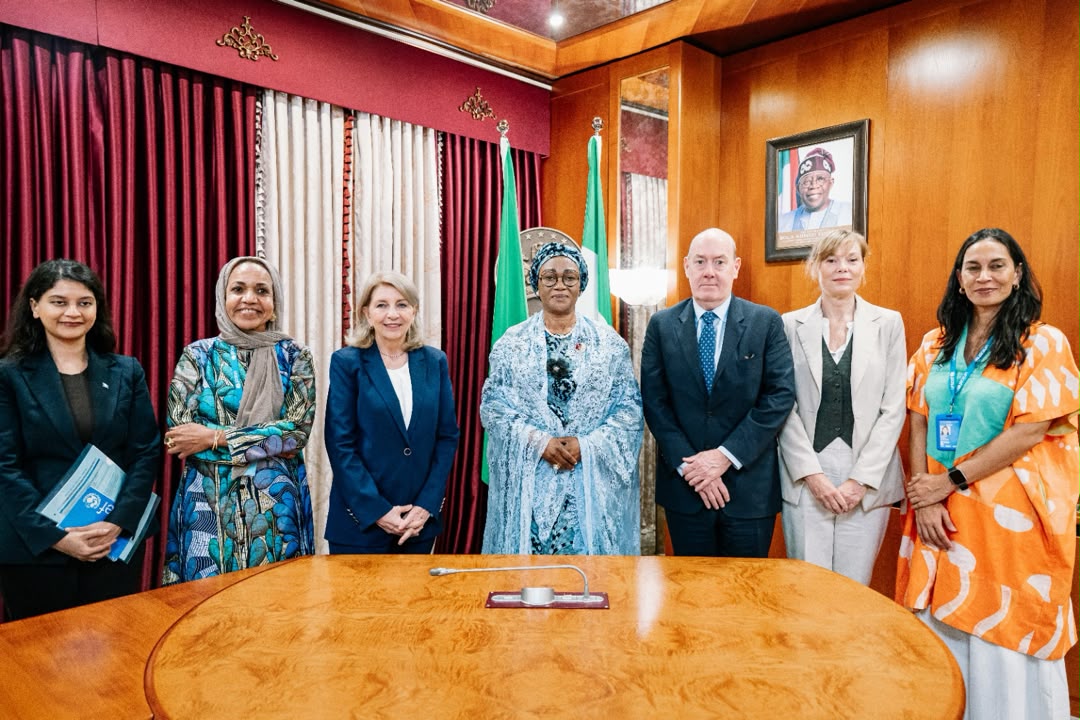 First Lady Senator Oluremi Tinubu, CON, wearing a light blue lace outfit and head wrap, standing center behind a conference table with a diverse group of delegates and officials indoors.